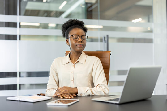 African American businesswoman meditating at desk in office. Woman practicing relaxation techniques during work day. Concept of stress management and mindfulness in professional environment.