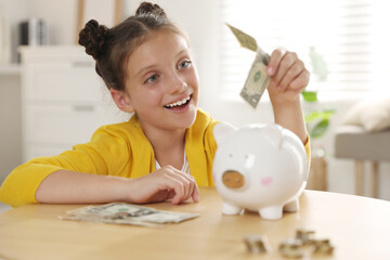 Pocket money. Cute girl putting dollar banknote into piggy bank at wooden table indoors