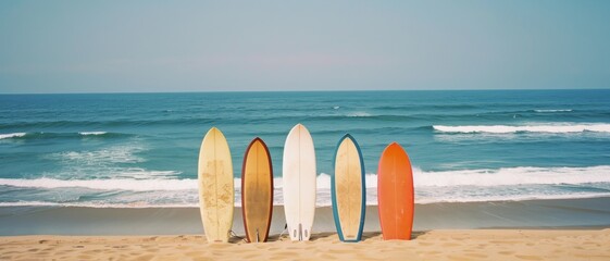 A set of five surfboards of different colors and styles stand upright on a sandy beach, with the ocean waves rolling in the background under a clear sky.