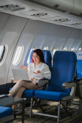 In-Flight Productivity: A young Asian businesswoman conducts a video call on her laptop while seated in an airplane cabin, showcasing the ease of modern business travel.