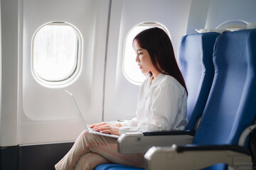 Fototapeta premium Businesswoman Working on Laptop During Flight: A young businesswoman in a white shirt and khaki pants is focused on her laptop while seated on an airplane. The image captures the essence of modern bus