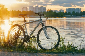 A bicycle resting against a riverbank during a golden sunset, with city buildings in the distant background, capturing a serene, urban outdoor moment.