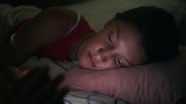 A teenage girl lies lazily on her bed, scrolling through social media on her phone. The room is calm and quiet, with the soft glow of the screen reflecting on her face.