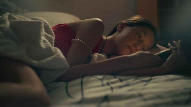 A teenage girl lies lazily on her bed, scrolling through social media on her phone. The room is calm and quiet, with the soft glow of the screen reflecting on her face.