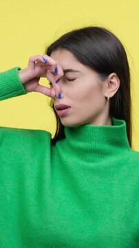 Dissatisfied Caucasian young adult woman covering nose from nasty or unpleasant smell, standing on yellow background. Vertical video.