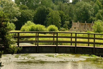 View of a wooden footbridge over River Coln to the Rack Isle water meadow and cottages in the Cotswold village of Bibury, England, UK