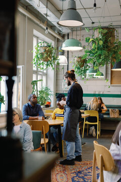 Male waiter taking order from couple near people resting in cafe
