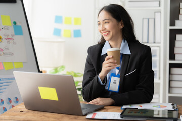 Confident and Composed: A professional woman in a business suit, a confident smile on her face, enjoys a cup of coffee while working on her laptop in a modern office setting. This image conveys a sens © Kritdanai