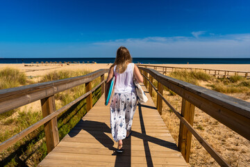 Long-haired woman wearing white top and white pants with colorful patterns walking to sandy beach and carrying bodyboard on sunny day on wooden promenade. Vacation in Algarve, Portugal. Back view