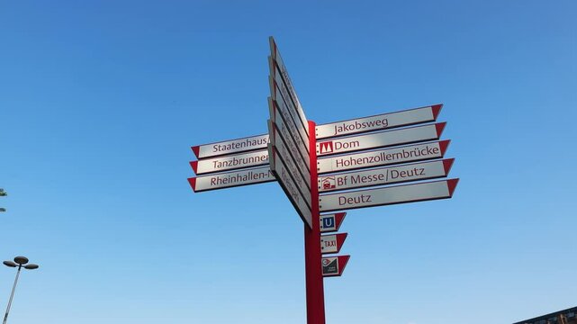 Direction sign with many different directions on it in Cologne, Germany. The sign is red and white. The sky is blue