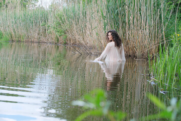 Sexy woman with curly hair in a sheer, flowing dress, serenely in a tranquil lake surrounded by...