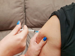 A man is getting a shot in his arm by a woman with blue nail polish, couch in the background. Amateur non professional Injection of medicine or vitamins due to poverty. Drug use and vaccination