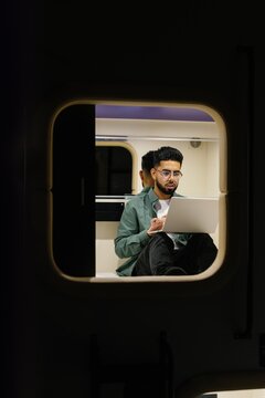 Young focused male student using laptop in economy-class capsule hotel