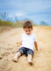 Young child playing happily in sandy terrain on a sunny day. A child in a white shirt enjoys sitting in the sand, smiling and playing under a blue sky on a warm day.