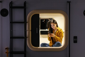 Asian woman messaging smartphone in mini-room of capsule hotel