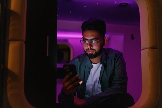 Man using smartphone in colorful mini-room of capsule hotel at night