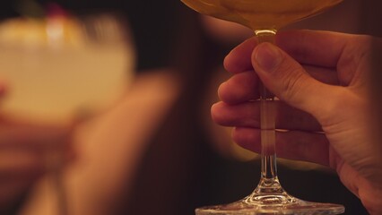 Closeup cropped woman hand making a toast mixed passion fruit smoothie cocktail wearing ring with friend at blurred drink background at nighttime warm light at nightclub counter bar party. Vinosity.