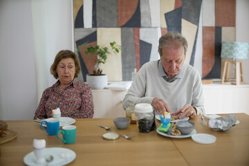 Elderly couple having breakfast at a wooden table with cups, jars, and plates, in a modern decorated dining room, showing a daily routine and the simplicity of shared meals together