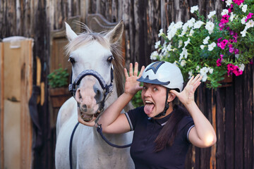 Woman rider in equestrian helmet, making funny face and sticking out tongue, horse near her also opening mouth