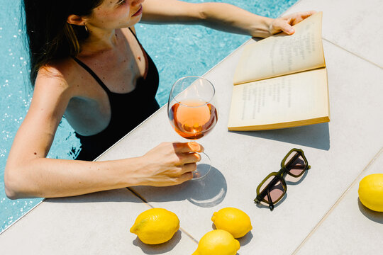 Lady relaxes by reading a book and drinking wine in a pool