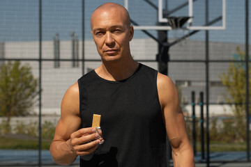 Sportsman eating energy bar. Muscular athlete snacks on protein bar while resting after workout at stadium. Healthy protein snack or energy bar, fitness dieting food for sporty lifestyle.