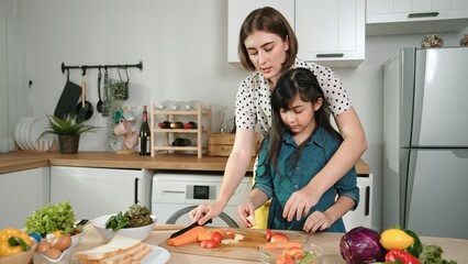 Smart caucasian mother and asian girl cooking together and chopping vegetable or preparing salad for dinner. Happy mom and daughter making healthy food with fresh food. Healthy food concept. Pedagogy.