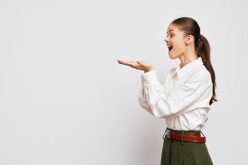 Young woman expressing excitement with hand gesture, wearing a white blouse and green pants, against a neutral gray background, showcasing a sense of surprise and joy