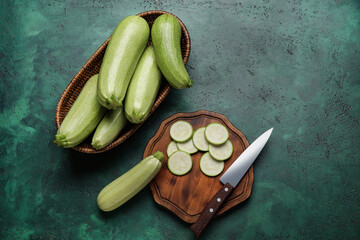 Wooden board and wicker basket with fresh zucchini on green background
