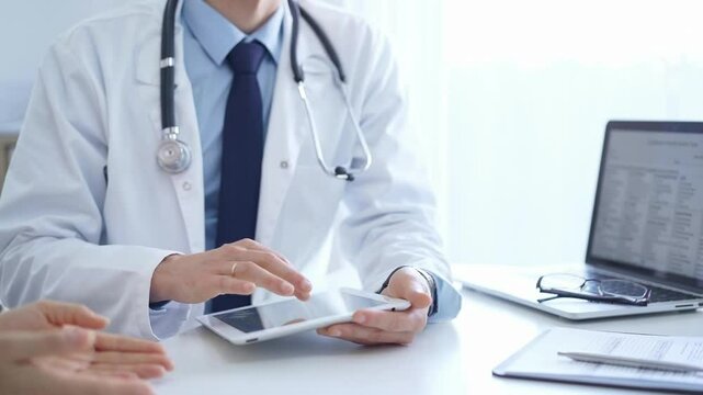 Doctor and a patient. The physician, wearing a white medical coat over a blue shirt and tie, is using tablet computer during a consultation in the clinic. Medicine