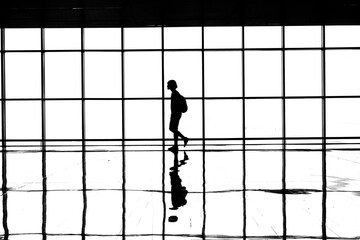 Young man with a backpack walking across reflective floor with multi paneled windows in the background, monochrome