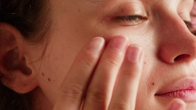 Young woman applying moisturizing cream on her face close-up. Morning skin care routine. Pink background