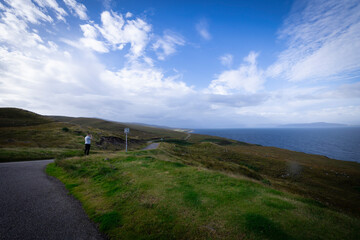 The image shows a person standing on a path by a body of water in Scotland. The surroundings include grass, clouds, sky, and other natural elements typical of the Scottish landscape.