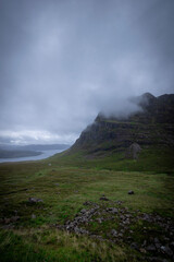 The image depicts a scenic road winding through misty mountains, with clouds hanging low in the sky. The landscape features lush greenery and the silhouette of hills in the background. scotland