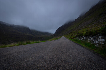 The image depicts a scenic road winding through misty mountains, with clouds hanging low in the sky. The landscape features lush greenery and the silhouette of hills in the background. scotland