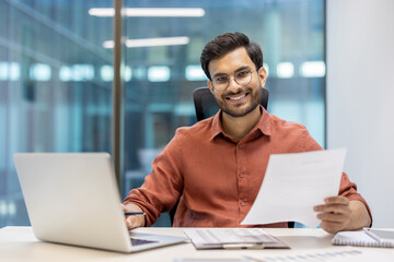 Smiling professional man in modern office holding document and working on laptop at desk