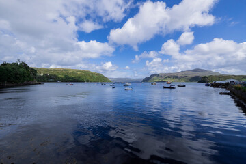 A serene landscape featuring a body of water with boats and buildings along the shore. The image captures a peaceful scene with a cloudy sky overhead.  Portree, Scotland, isle of skye