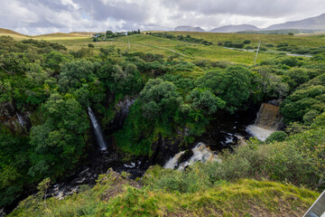 A stunning waterfall cascading down rocks in a lush green forest setting. The image captures the beauty of nature with a cloudy sky above. 