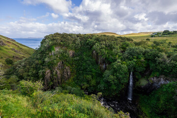 A stunning waterfall cascading down rocks in a lush green forest setting. The image captures the beauty of nature with a cloudy sky above. 