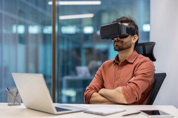Young professional wearing virtual reality headset sitting at office desk using innovative...