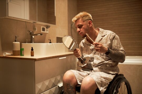 Person sitting in wheelchair grooming themselves in bathroom with a mirror. Bathroom details like sink, mirror, and essential grooming products are visible