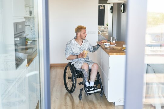 Person eating breakfast while sitting in wheelchair in modern kitchen setting with wooden floor. Holding beverage in stylish and accessible kitchen environment