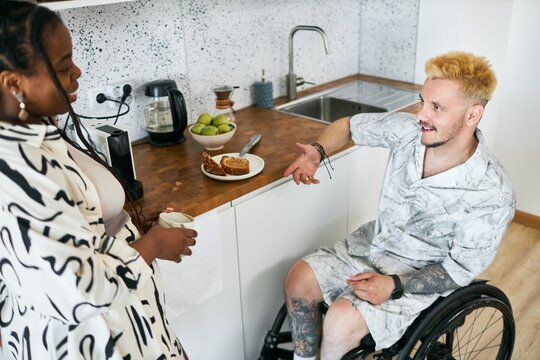 Man in wheelchair having friendly conversation with woman in contemporary kitchen while preparing meal and using devices