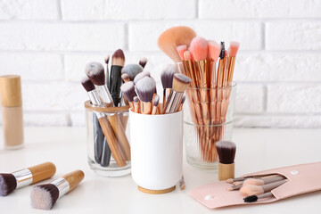 Holders with makeup brushes on table near white brick wall