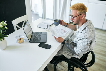 Man in wheelchair reading documents and holding mug while working at home office table showing...