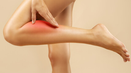 Sick or injured woman experiencing calf muscle pain standing on beige background, woman's leg with injury, sore spot is marked in red © Home-stock