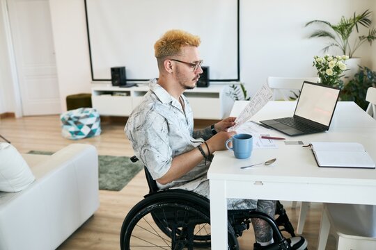 Man sitting in wheelchair at table reading business documents with laptop and coffee mug present Room elegantly decorated with flowers and modern accessories surrounding