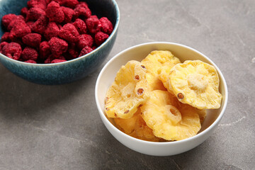 Bowls of dehydrated raspberries and pineapple on grey background