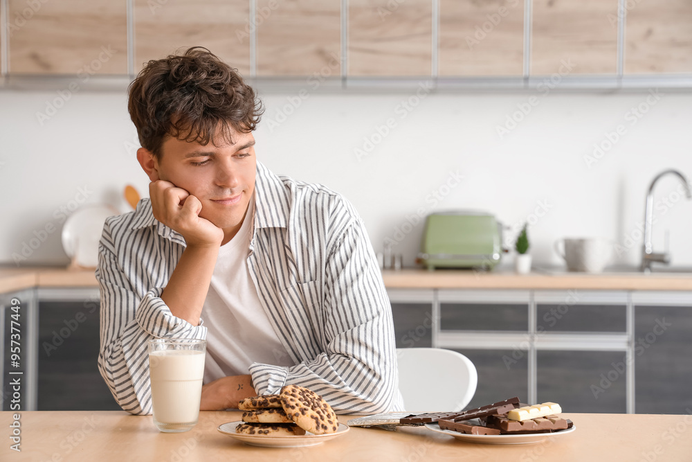 Sticker Handsome young man with plate of sweet chocolate bars, tasty cookies and glass of milk at table in kitchen