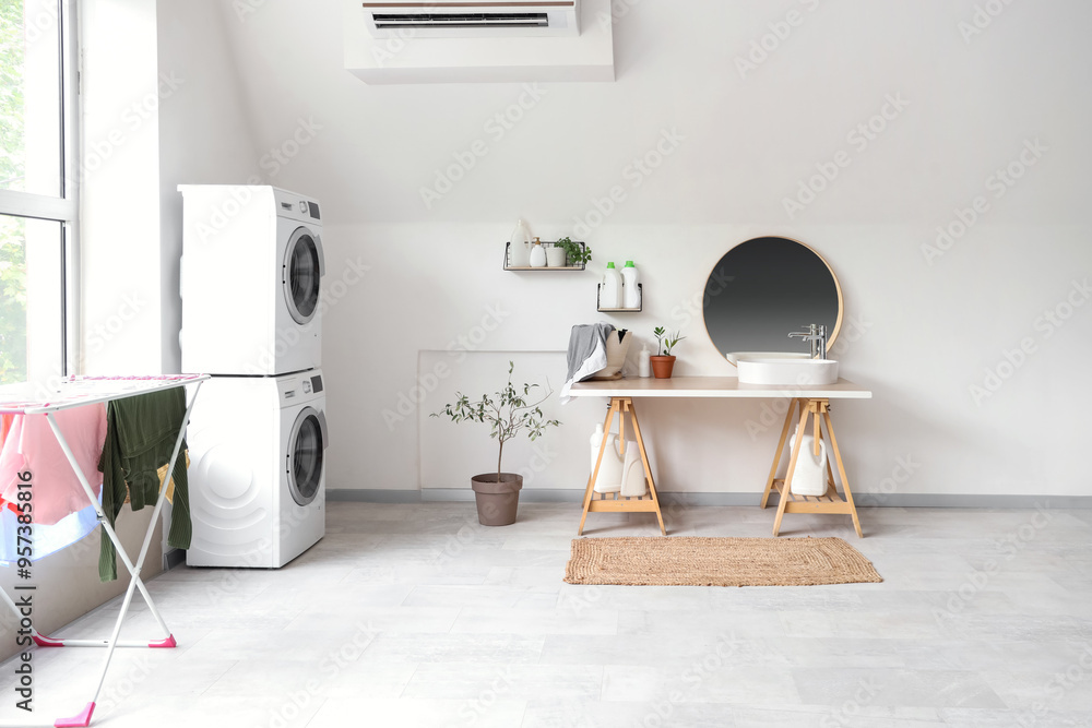 Poster Interior of light bathroom with washing machines, dryer, sink and laundry basket on table