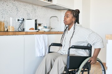 An African American woman with box braids sitting in wheelchair in modern kitchen, looking to the side. Countertops with neatly organized items is visible in the background
